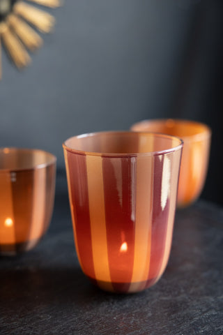 The Deep Bordeaux Striped Tealight Holder - Tall with a lit tealight inside, displayed on a dark surface with two other tealight holders in the background.