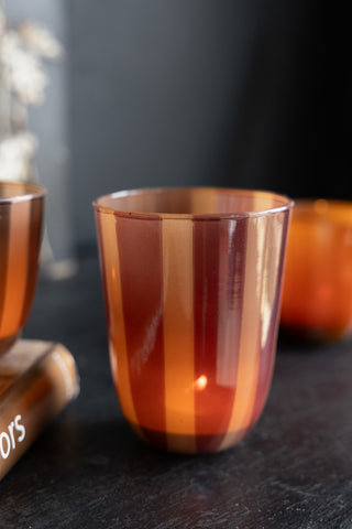 Close-up of the Deep Bordeaux Striped Tealight Holder - Tall with a lit tealight inside, styled on a dark surface with a book and other small tealight holders.