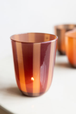 The Deep Bordeaux Striped Tealight Holder - Tall with a lit tealight inside, on a table with other tealight holders in the background.