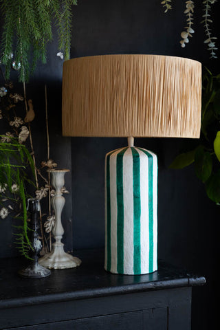 The Deep Green & Off White Stripe Paper Mache Table Lamp styled on a sideboard with two candlestick holders and some greenery.