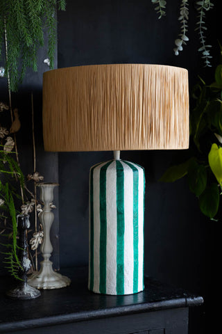 The Deep Green & Off White Stripe Paper Mache Table Lamp displayed on a black sideboard with some candlestick holders, surrounded by greenery.