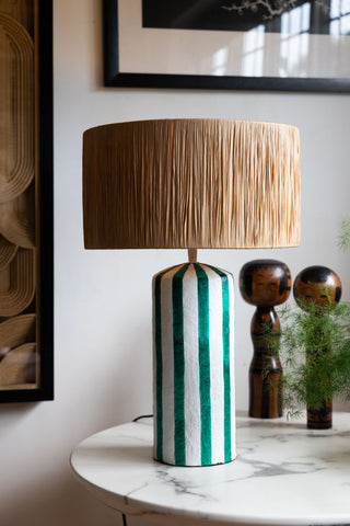 The Deep Green & Off White Stripe Paper Mache Table Lamp styled on a round table with a plant and some ornaments.