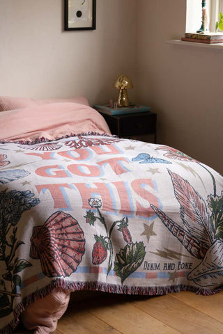 The Denim & Bone You Got This Woven Cotton Throw styled on a bed in a bedroom, with a bedside table and various home accessories.