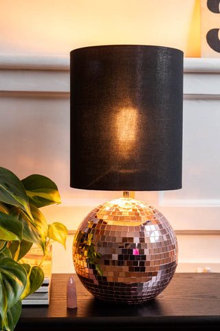 The Disco Ball Table Lamp in Pink switched on, styled on a dark surface with some magazines, a plant and a pink crystal.