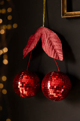 The Disco Cherries Christmas Tree Decoration hanging in front of a dark wall with a metallic frame and some fairy lights in the background.