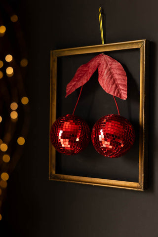 The Disco Cherries Christmas Tree Decoration hanging on a dark wall inside a frame, with fairy lights in the background of the shot.