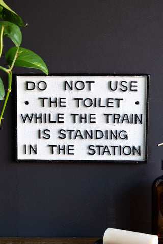 The Do Not Use The Toilet When The Train... Hanging Sign displayed on a black wall, styled with some greenery, a soap dispenser and a bar of soap.