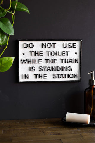 The Do Not Use The Toilet When The Train... Hanging Sign displayed on a black wall, styled above a wooden surface with a soap dispenser, bar of soap on a soap dish and some greenery.