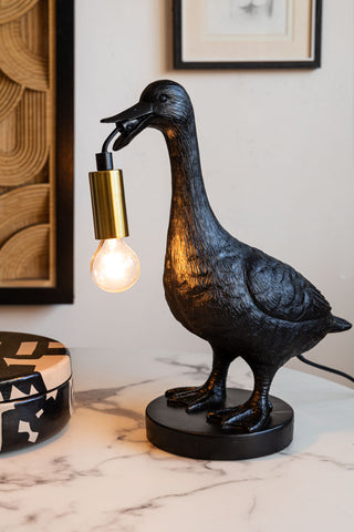 The Duck Table Lamp switched on and styled on a table with a trinket box, with artwork on the wall in the background.