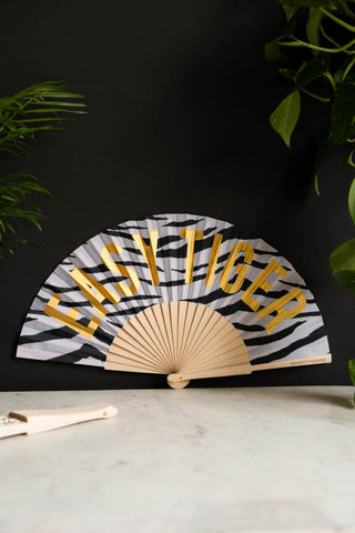 The Easy Tiger Print Fan displayed on a marble table leaning against a black wall, styled with another fan and some plants.