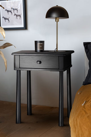 The Elegant Black Bedside Table styled next to a bed, with a candle and a lamp on.