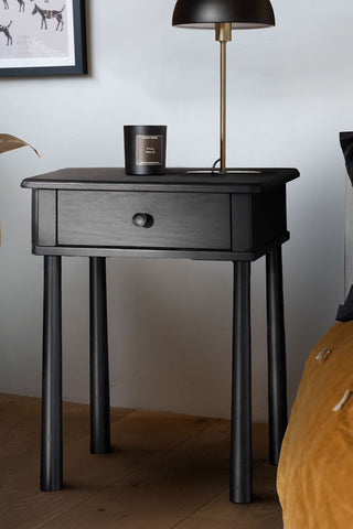 The Elegant Black Bedside Table next to a bed, styled with a candle and a table lamp on. 