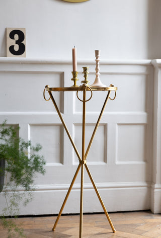 The Elegant Gold & Glass Side Table in front of a white panelled wall, styled with candlestick holders on top and a potted plant and piece of art in the background.