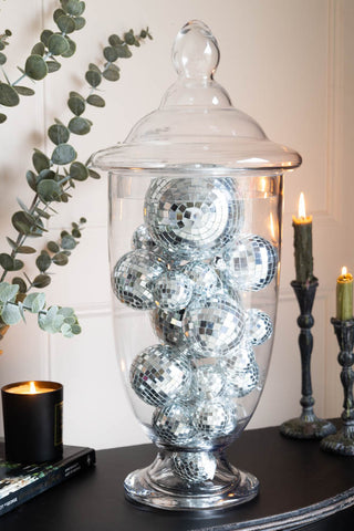 Large decorative glass jar with a smooth finish and lid filled with silver disco balls, set on a black console table with glowing candles and eucalyptus.