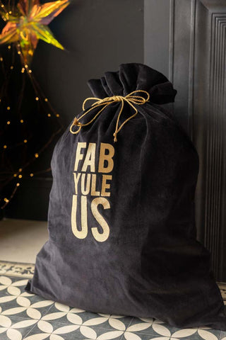 The Fab Yule Us Velvet Christmas Sack styled on a geometric floor in front of a black storage unit, with a star decoration and some fairy lights in the background.