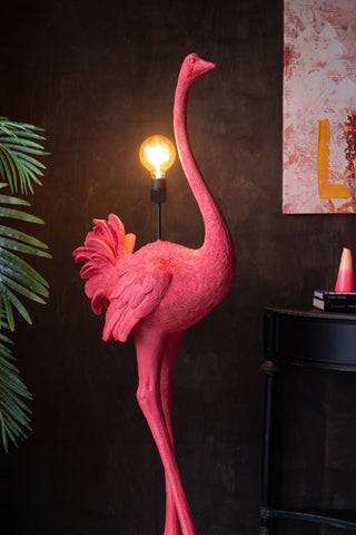 The Fabulous Pink Ostrich Floor Lamp switched on and styled in front of a dark wall, next to a plant, table with accessories and artwork on the wall.