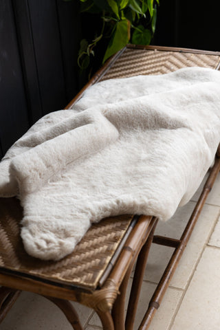 The Faux Fur Natural Rug displayed draped across a wicker bench, with some greenery in the background.