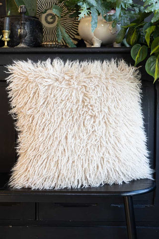 The Faux Mongolian Natural Cushion displayed on a black bench in front of a sideboard styled with home accessories and greenery.