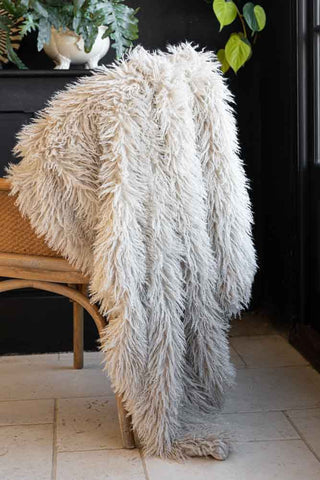 The Faux Mongolian Natural Throw displayed draped across a wicker chair in the corner of a room, styled with some plants.