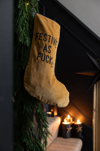 The Luxury Festive As Fuck Gold Christmas Stocking displayed on some stairs with a garland, with lit candles and a star decoration in the background.