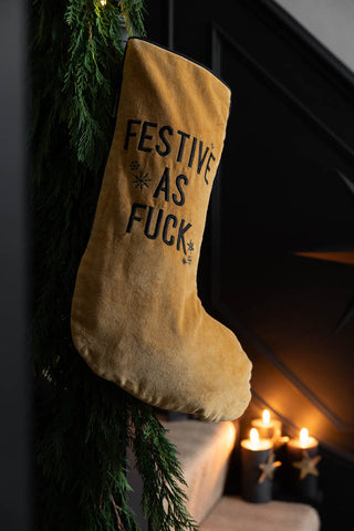 The Luxury Festive As Fuck Gold Christmas Stocking hanging on some stairs with a garland, with lit candles in the background.