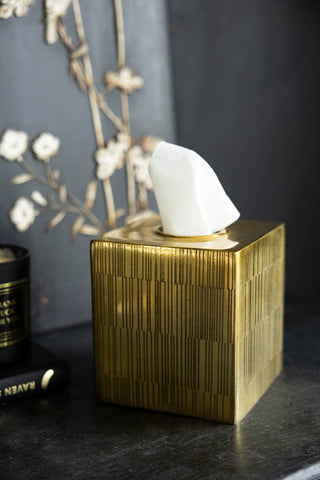 The Fine Line Detail Gold Effect Tissue Box with tissues inside, displayed on a black sideboard with a candle and a book with a floral decoration in the background.