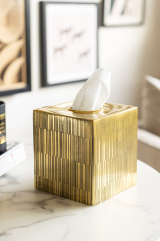 The Fine Line Detail Gold Effect Tissue Box with tissues inside, styled on a white marble table with some other home accessories, with a sofa and some art prints in the background.