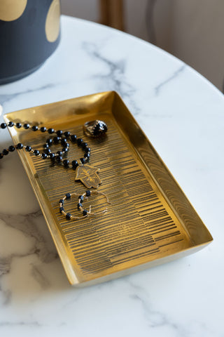 The Fine Line Detail Gold Effect Trinket Tray styled with jewellery inside, displayed on a white marble table with a black and gold pot in the background.