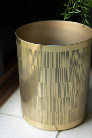 Close-up of the Fine Line Detail Gold Effect Waste Bin with the leaves of a plant in the background.