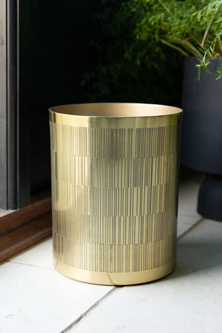 The Fine Line Detail Gold Effect Waste Bin displayed on the floor next to a door frame and a potted plant. 