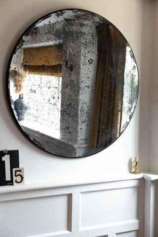 A round antique-style mirror on a white wall above some decorative numbers and a candle in a candlestick holder.