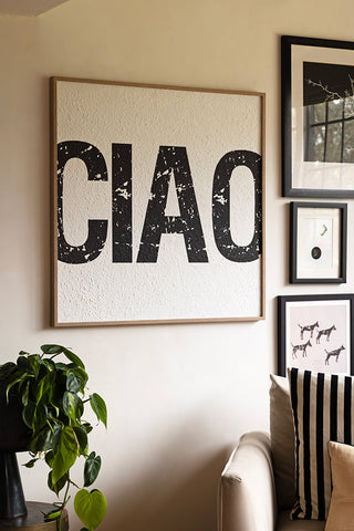 The Framed Black & White Ciao Wall Art styled on the wall with some other art prints, displayed next to a potted plant and a sofa with some cushions on.