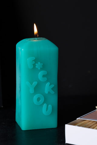 Close-up of the Fuck You Candle lit, displayed on a dark surface with a box of matches.