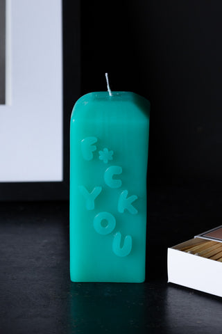 The Fuck You Candle displayed on a dark surface next to a box of matches and a framed artwork.