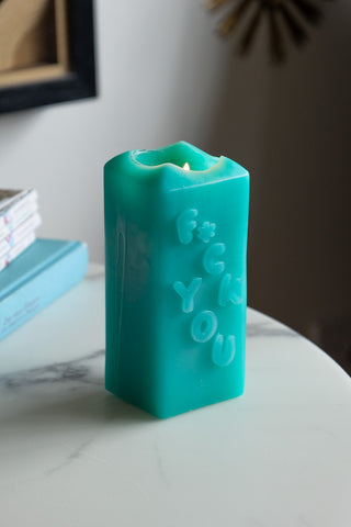 The Fuck You Candle lit, displayed on a white marble surface with some books in the background.