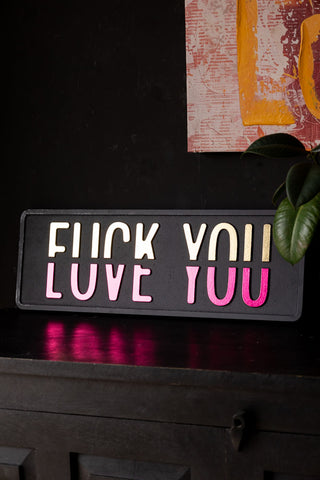 The Fuck vs Love You Wall Sign styled on a sideboard, leaning against a wall with another piece of artwork on, next to a plant.