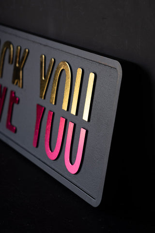 Close-up of the Fuck vs Love You Wall Sign, seen from an angle leaning against a black wall.