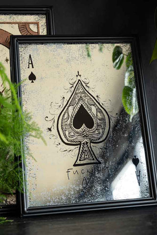 The Fucking Ace Decorative Playing Card Mirror leaning against a wall with another mirror, styled with some greenery.