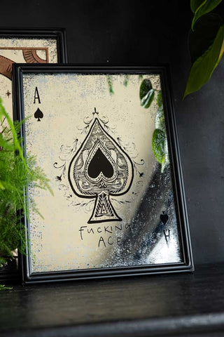 The Fucking Ace Decorative Playing Card Mirror styled on a black sideboard with another mirror and some greenery.