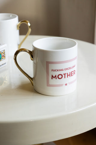 A white ceramic statement mug, featuring a metallic gold handle and red detailing 