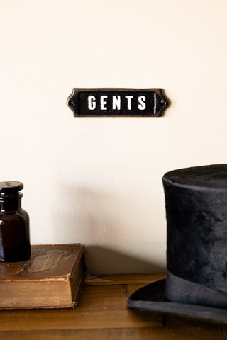 The Gents Door Hanging Sign displayed on a neutral wall above a wooden desk, styled with a top hat, book and glass bottle.