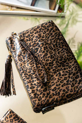 The Glam Leopard Print Clutch Bag displayed on a surface with a matching card holder, some magazines and some greenery.