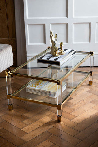 The Gold & Glass Coffee Table styled with various home accessories and magazines on, displayed on a wooden floor.