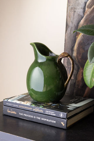 The Glazed Jug in Olive Green displayed on some books on a black sideboard, with a plant and artwork.