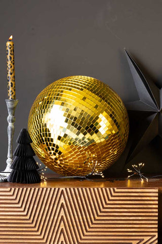 The Gold Disco Ball - 30cm styled on a wooden unit with a candlestick holder, fairy lights and paper Christmas decorations.