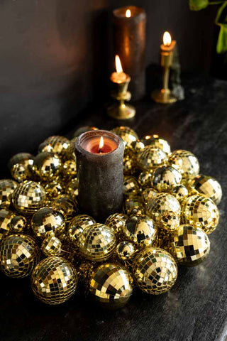 The Gold Disco Ball Wreath styled on a black sideboard with a lit black candle inside, with other lit candles/candlestick holders and a plant in the background.