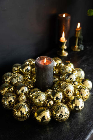 The Gold Disco Ball Wreath displayed on a black sideboard with a lit candle in the centre and other lit candles in the background of the shot.