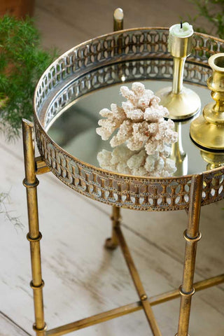 The Gold Distressed Mirrored Side Table styled with accessories on and a plant in the background.
