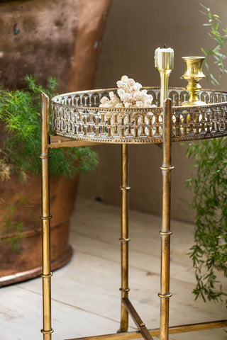 The Gold Distressed Mirrored Side Table in a bathroom styled with various accessories on, and a plant and metal bath in the background.