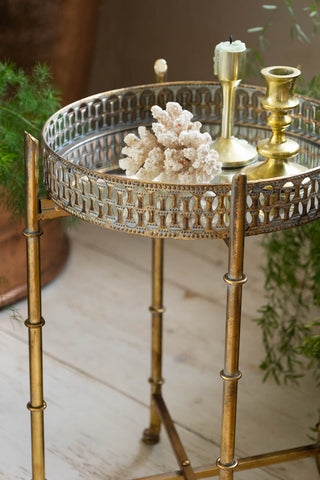 Detail shot of the Gold Distressed Mirrored Side Table, styled with various accessories on and plants to each side.